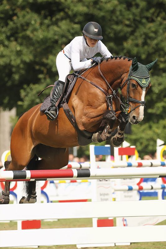 Eine Reiterin in weißer Kleidung springt mit ihrem braunen Pferd über ein Hindernis.