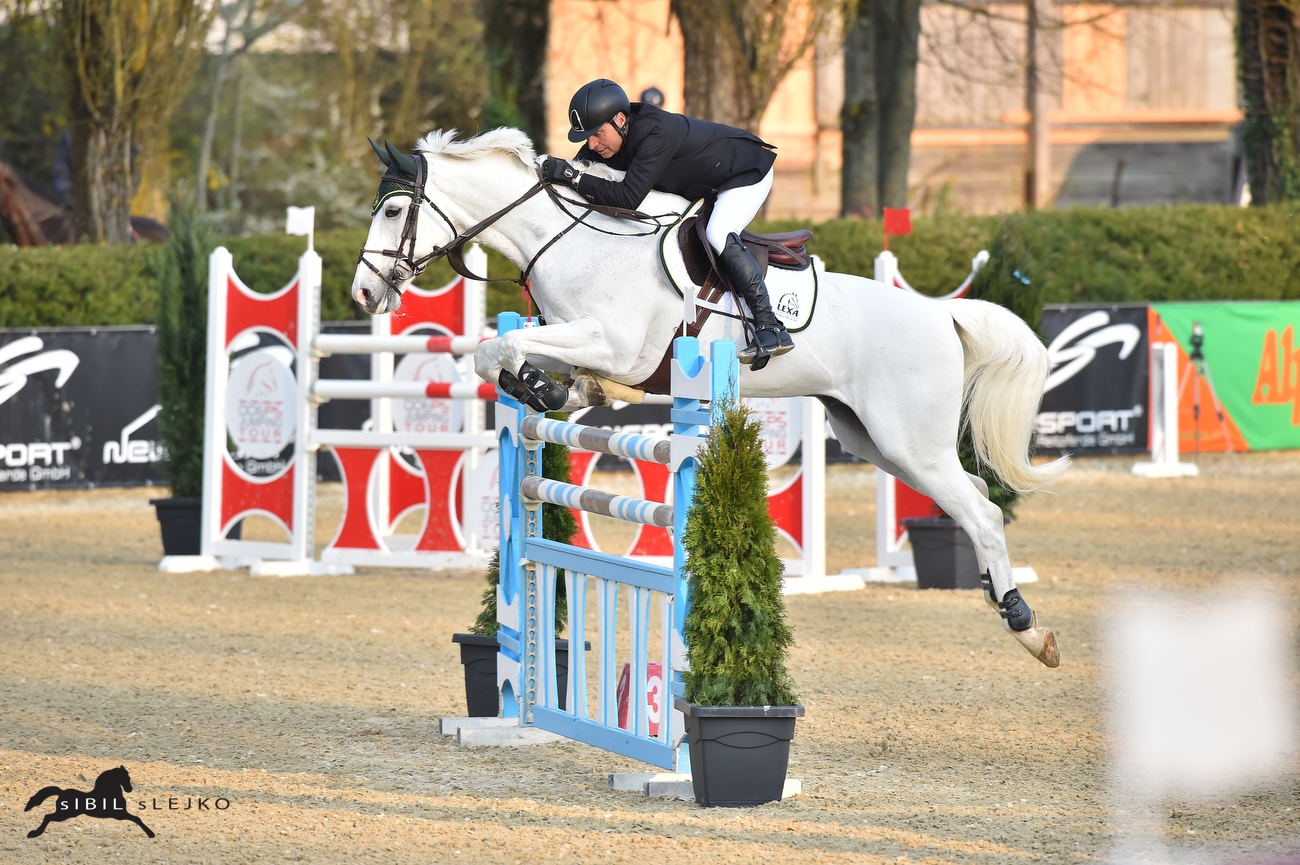 Ein Reiter in schwarzer Kleidung springt mit einem weißen Pferd über ein blaues Hindernis in einem Springturnier.