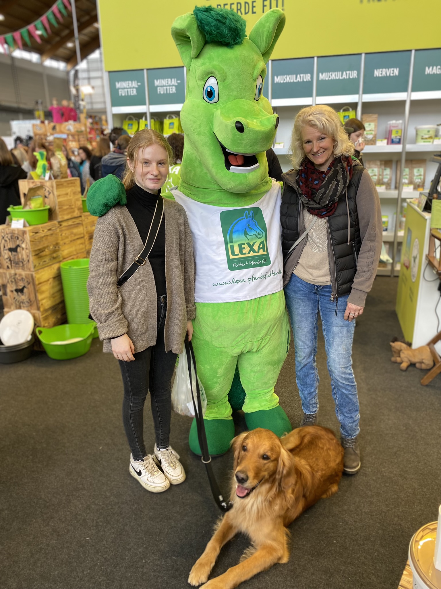 Eine Person in einem grünen Pferdekostüm steht neben zwei Frauen, eine mit Hund, auf einer Messe.
