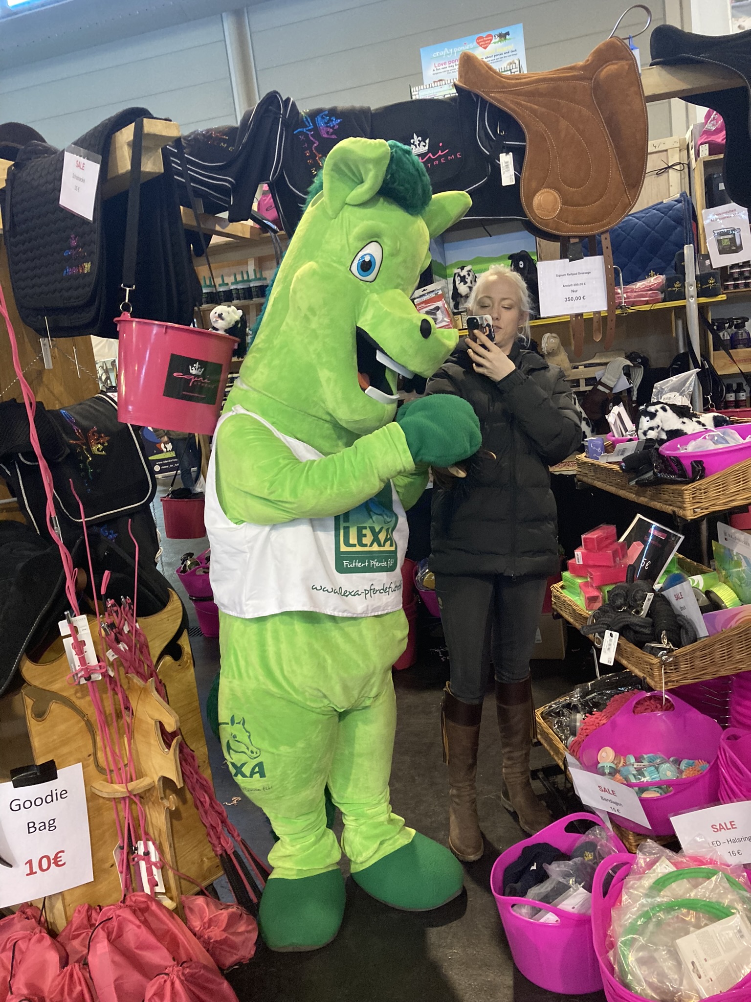 Ein grünes Maskottchen in einem Geschäft, das mit einer Person posiert, die ein Smartphone hält. Umgeben von verschiedenen Produkten.