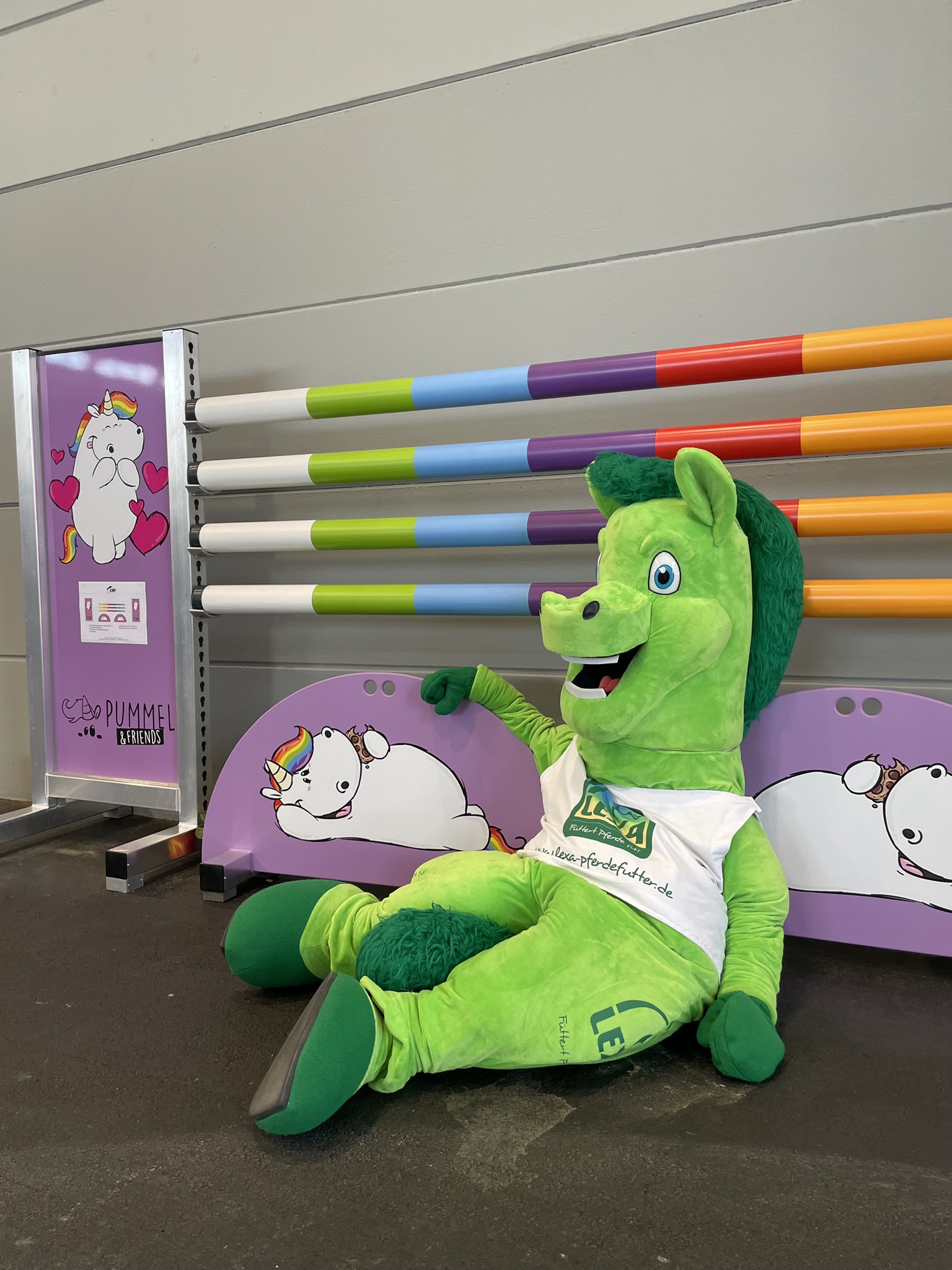 Ein grünes Maskottchen mit Pferdeform sitzt entspannt auf dem Boden, umgeben von bunten Stangen und einem Hintergrund mit Einhornmotiven.