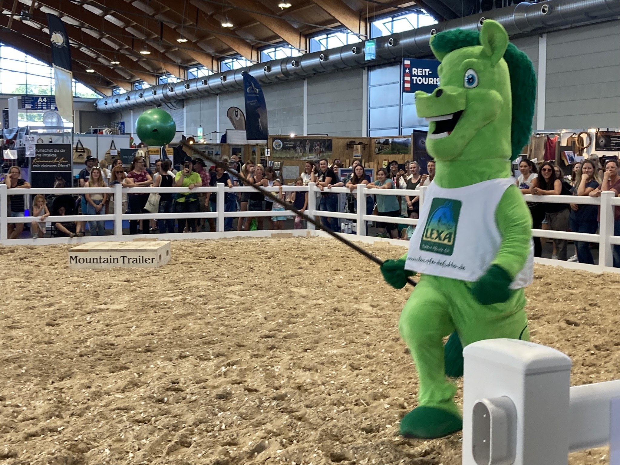 Ein grünes Maskottchen in einem Reitstall, das mit einem Stock einen Ball hält, umgeben von Zuschauern.