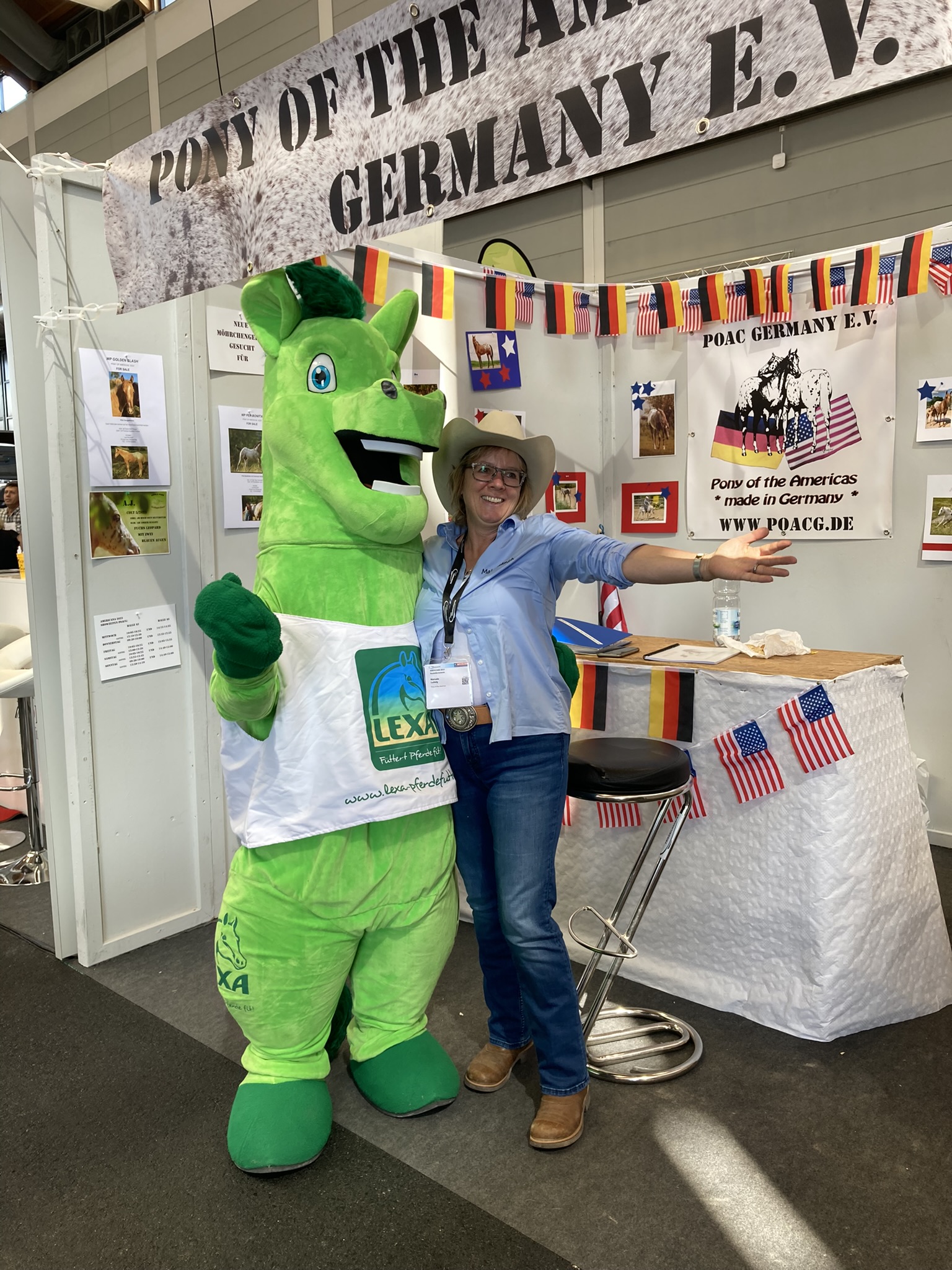 Eine Frau in einem Cowboyhut steht neben einem grünen Maskottchen mit einem Lächeln. Im Hintergrund sind Flaggen und ein Informationsstand zu sehen.