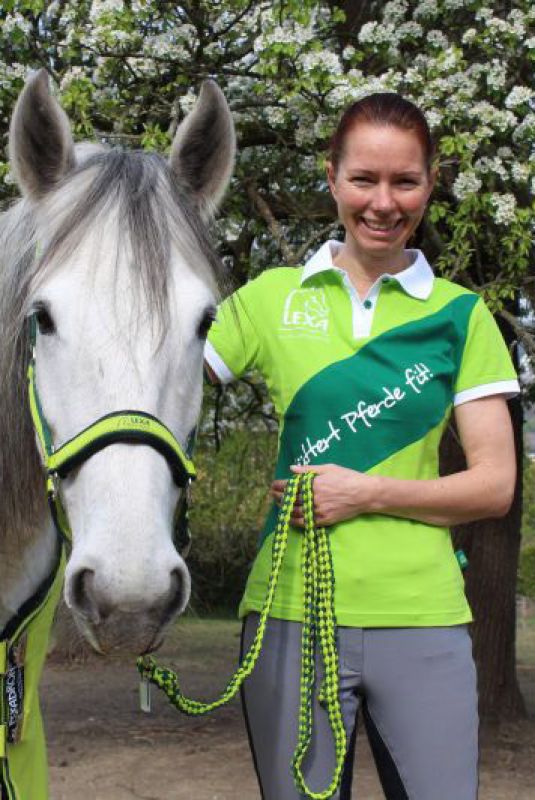Eine Frau mit langen roten Haaren steht neben einem grauen Pferd und hält dessen Zügel. Sie trägt ein grünes Polo-Shirt mit einem Logo.