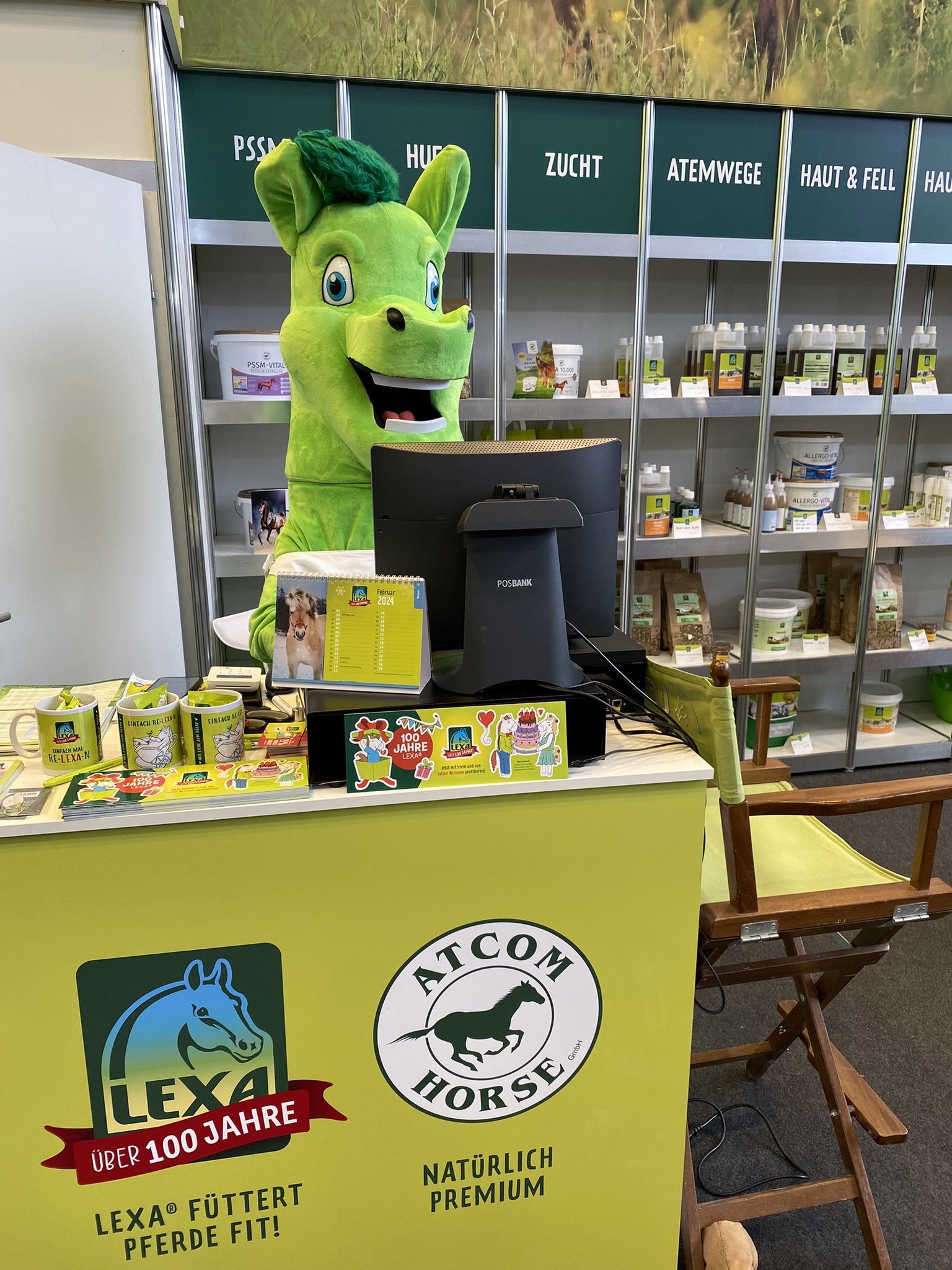Ein grünes Maskottchen steht an einem Tisch mit Produkten von LEXA und Atcom Horse, im Hintergrund Regale mit weiteren Produkten.