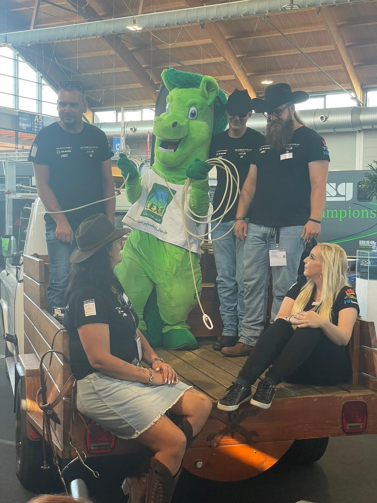 Eine grüne Maskottchenfigur steht auf einem Pickup, umgeben von mehreren Personen in Cowboyhüten und T-Shirts.