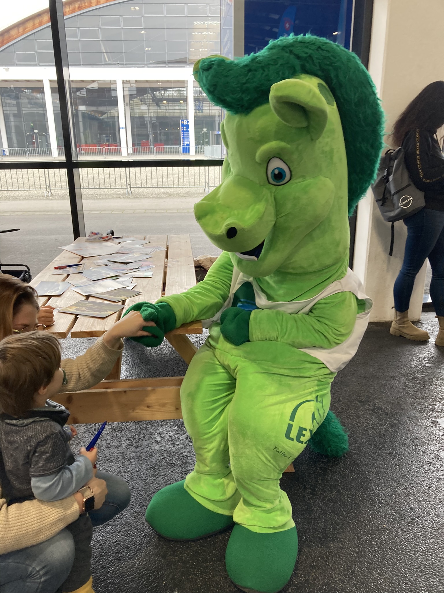 Ein grünes Maskottchen mit einem freundlichen Gesicht sitzt auf einer Bank und interagiert mit einem Kind.