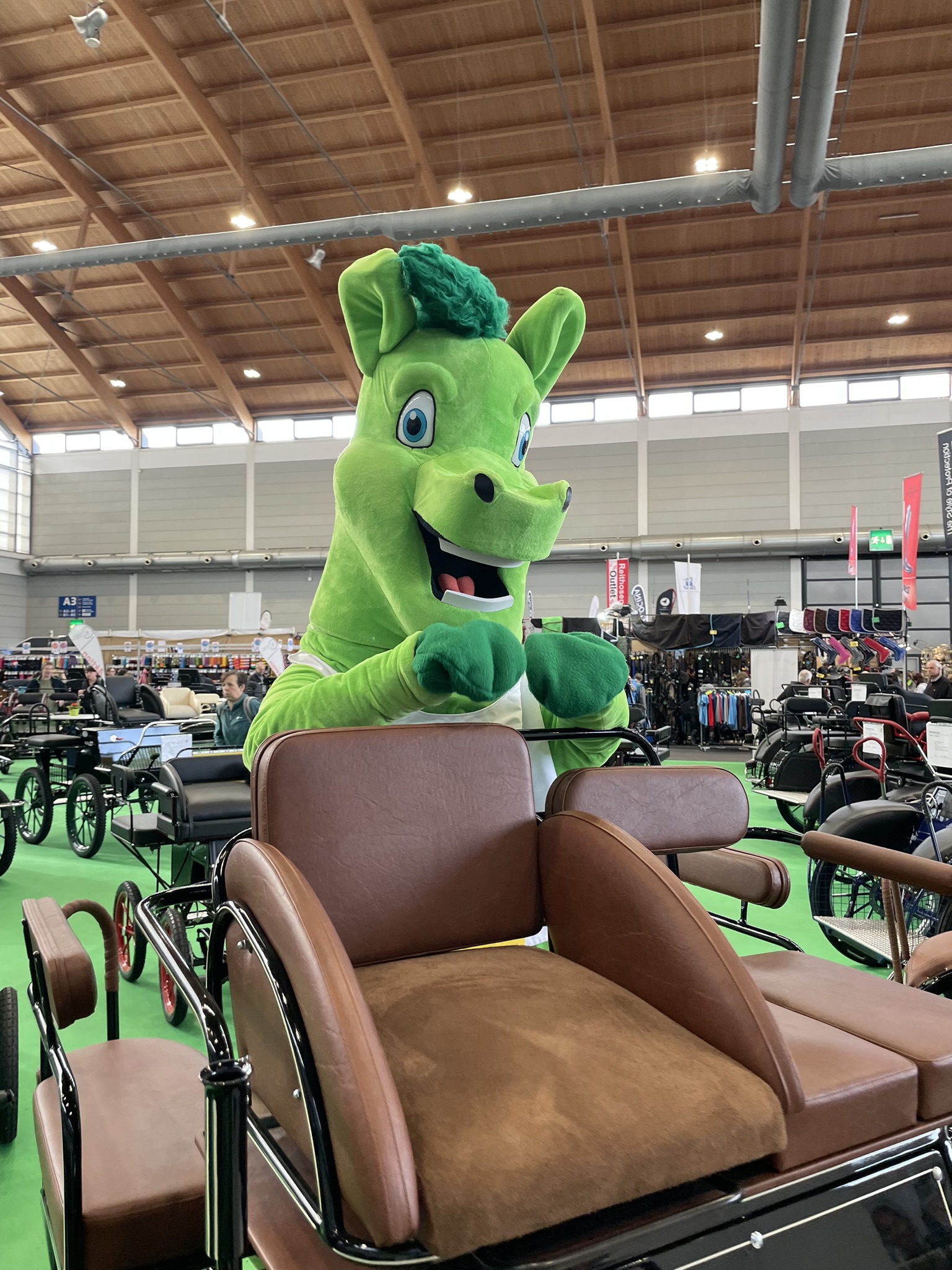 Ein grünes Maskottchen in Form eines Pferdes steht hinter einem Wagen mit braunen Sitzen.