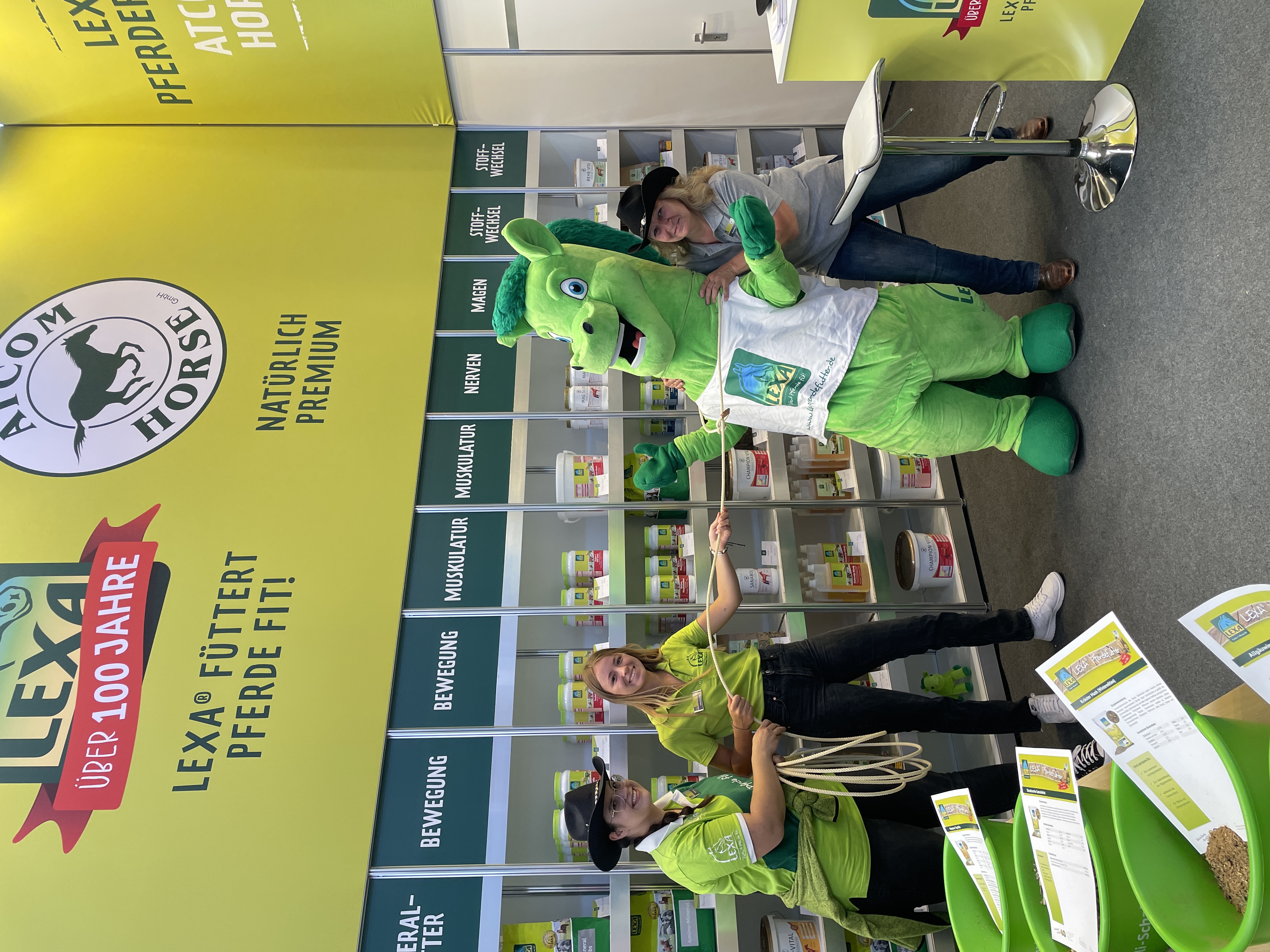 Grüne Pferde-Maskottchen steht neben zwei Personen in grünen Shirts an einem Messestand mit Produkten für Pferde.
