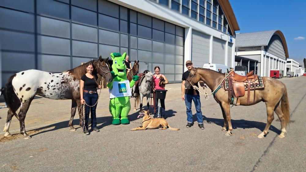 Eine Gruppe von Menschen steht mit zwei Pferden und einem Hund vor einem großen Gebäude. Eine Person trägt ein grünes Maskottchen-Kostüm.