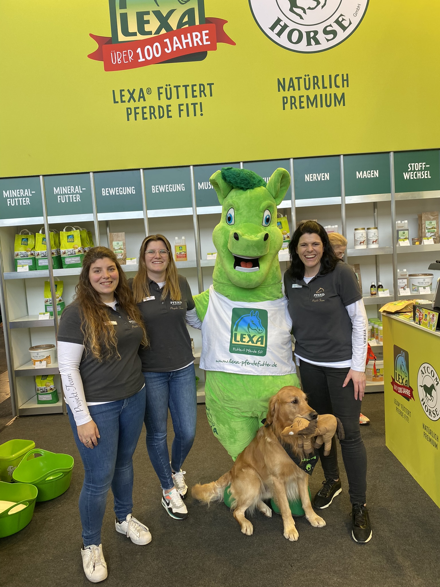 Drei Frauen stehen mit einem Maskottchen und einem Hund vor einem Stand von LEXA, der Produkte für Pferde anbietet.