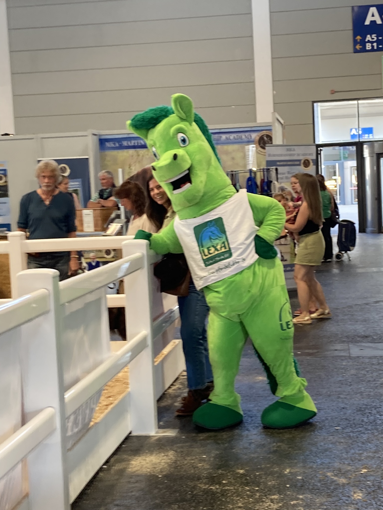 Ein grüner Pferdemaskottchen steht an einem Zaun, umgeben von Menschen auf einer Messe.