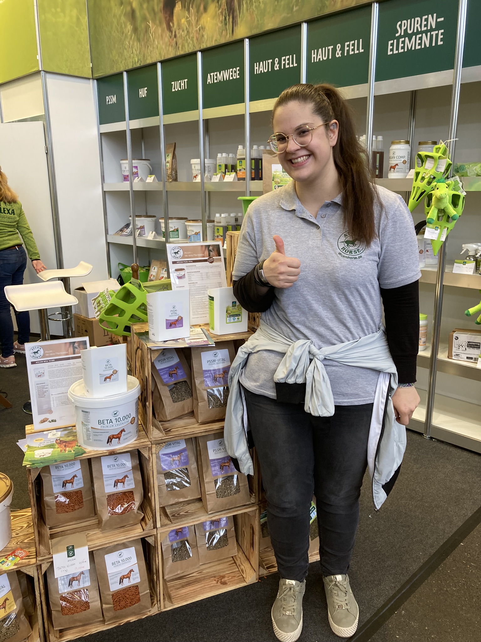 Frau mit Brille und grauem T-Shirt gibt Daumen hoch neben einem Stand mit Tierprodukten.