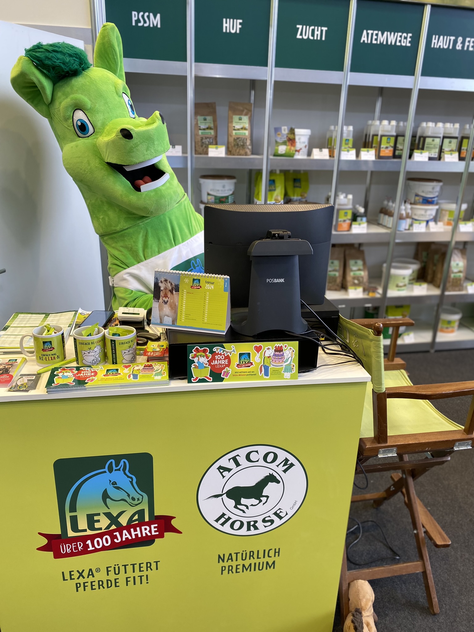 Ein grünes Maskottchen mit Pferdekopf steht an einem Tisch mit Produkten von LEXA und ATCOM HORSE im Hintergrund.