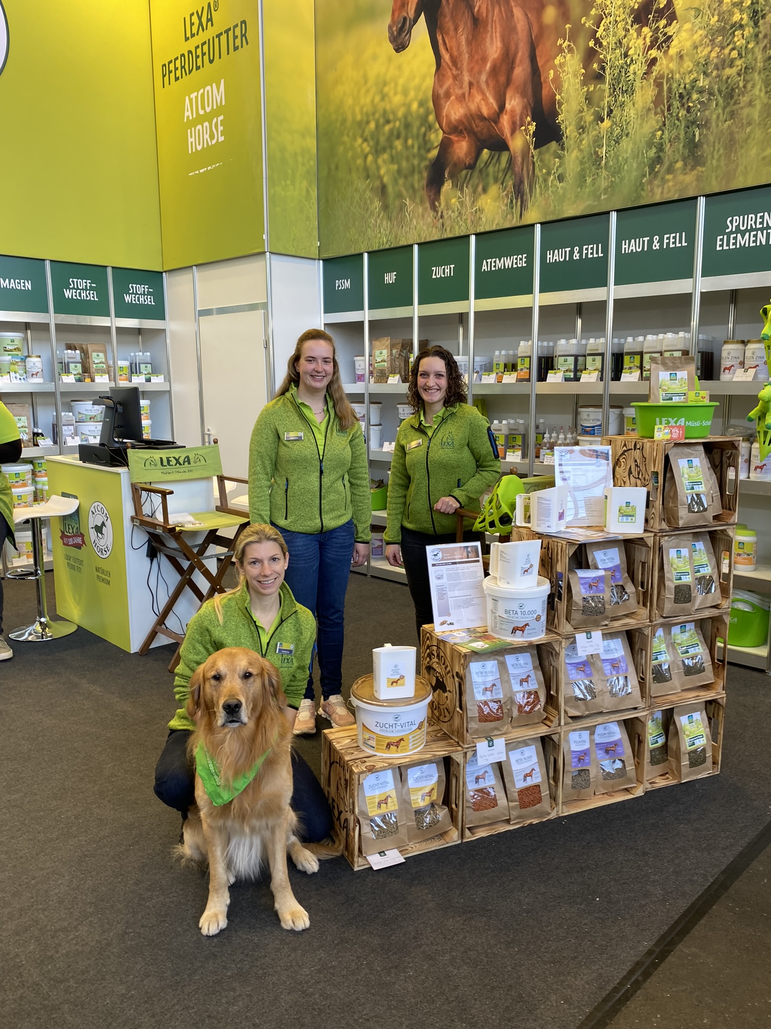 Drei Frauen in grünen Jacken stehen neben einem Hund vor einem Stand mit Tierfutterprodukten.