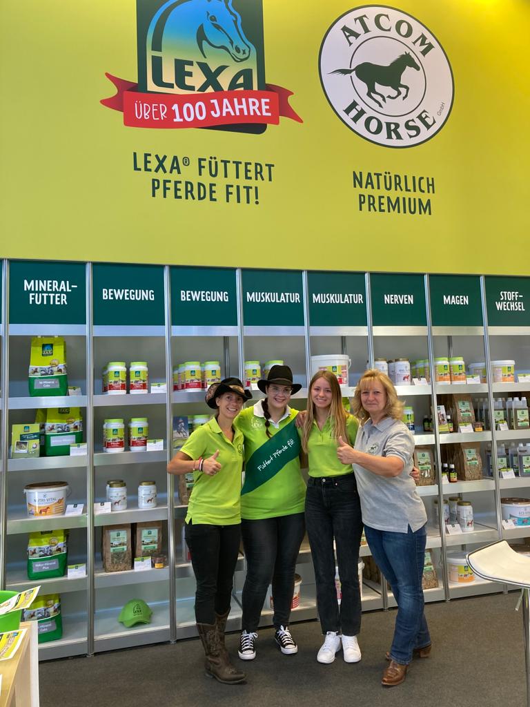Drei Personen in grünen T-Shirts stehen vor einem Regal mit Pferdefutterprodukten. Hintergrund mit dem Slogan 'LEXA® füttert Pferde fit!'