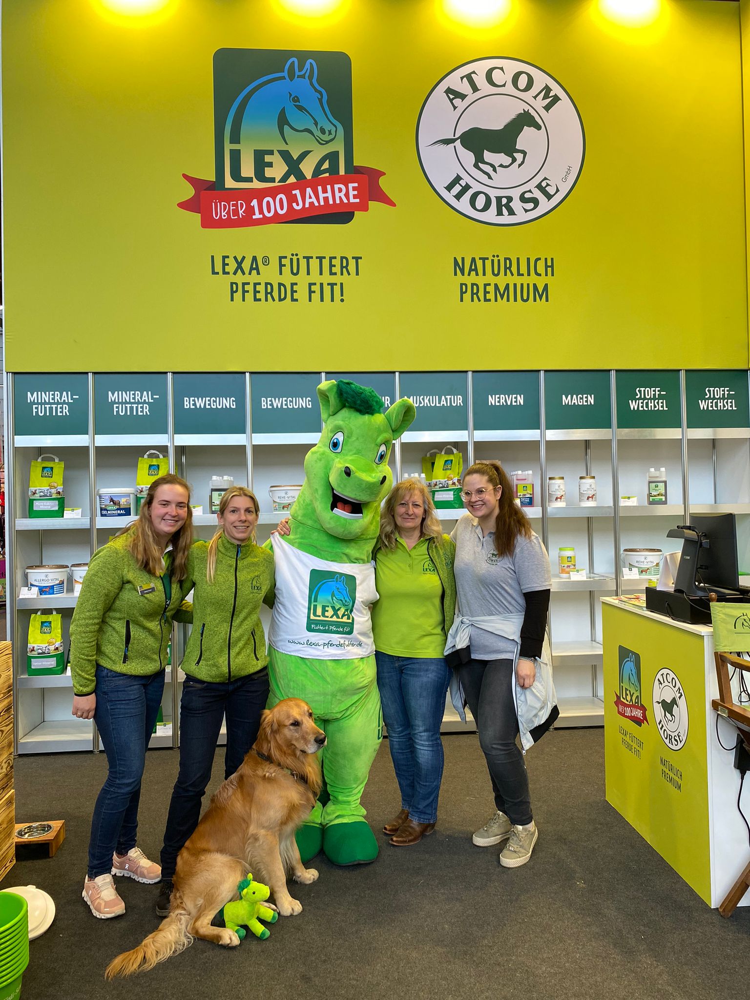 Gruppe von vier Personen und einem Hund, posierend mit einem Maskottchen in einem Stand für Pferdefutter.