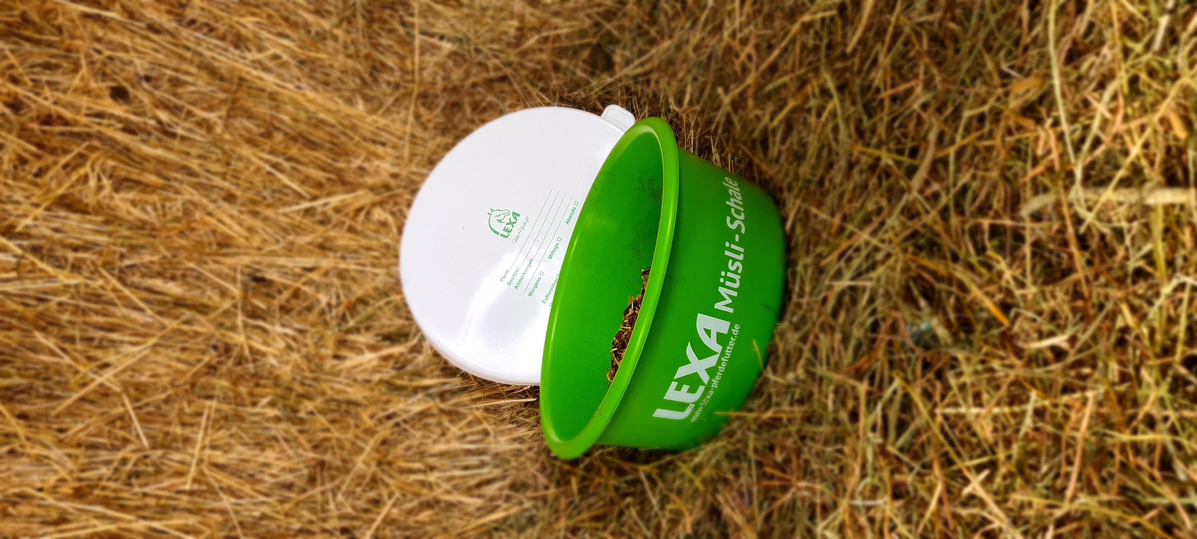 Grüner Eimer mit der Aufschrift 'LEXA Müsli-Schale', gefüllt mit Müsli, auf trockenem Gras.