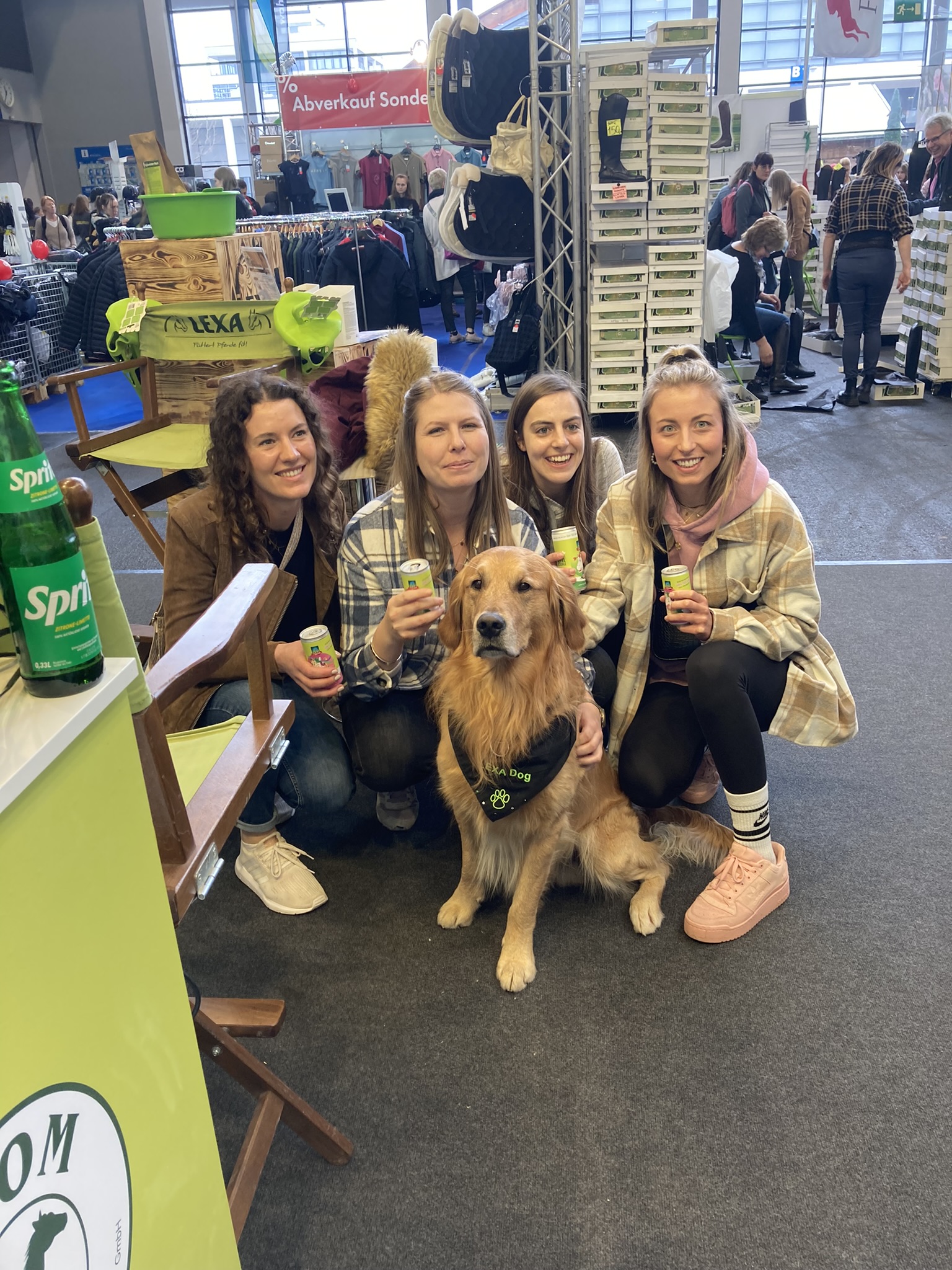 Fünf Personen, darunter ein Golden Retriever, posieren lächelnd in einem Ausstellungsraum. Alle halten Getränke in der Hand.