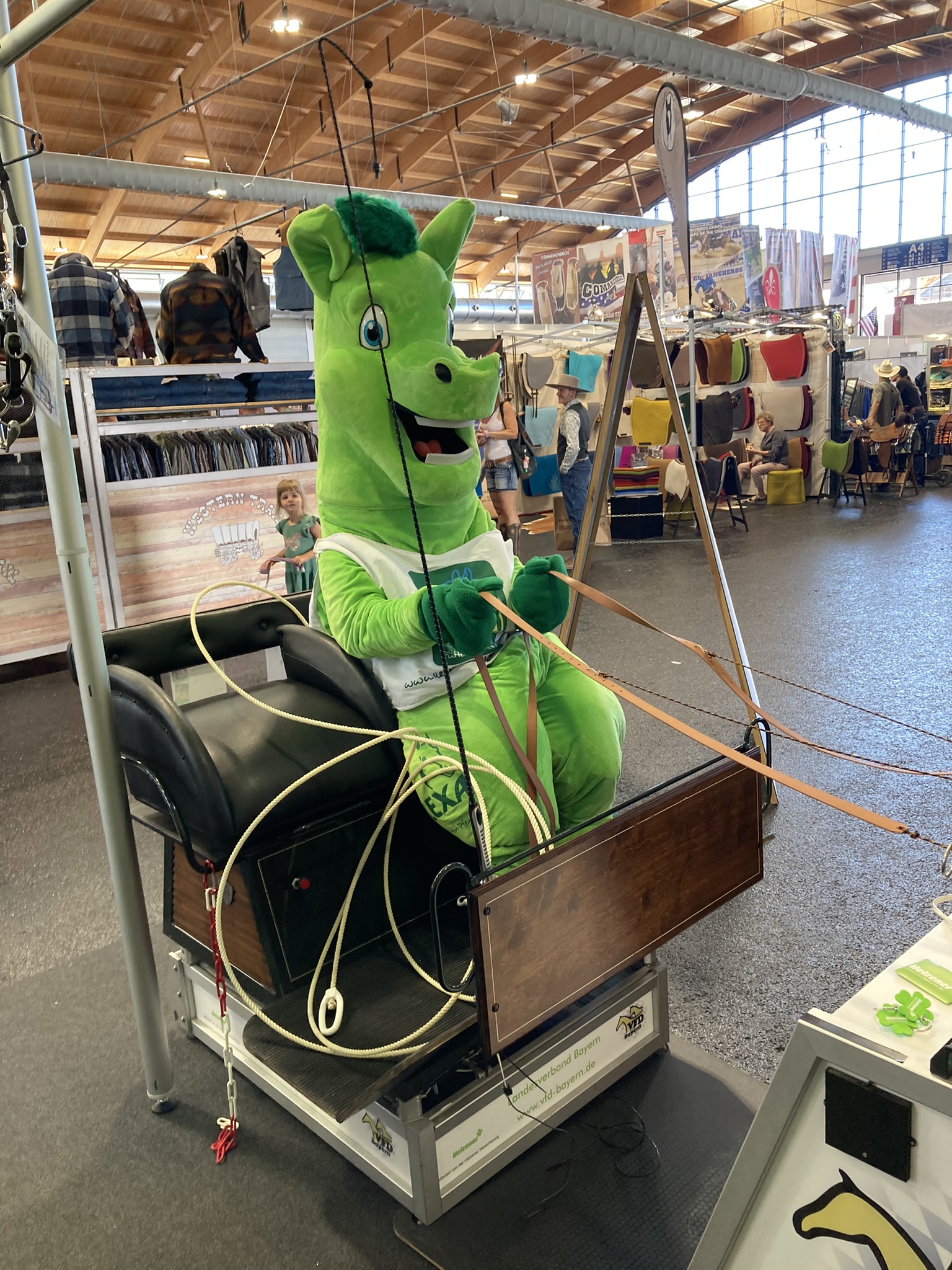 Ein grünes Maskottchen in Form eines Drachen sitzt auf einem Sattel und ist mit Seilen verbunden.