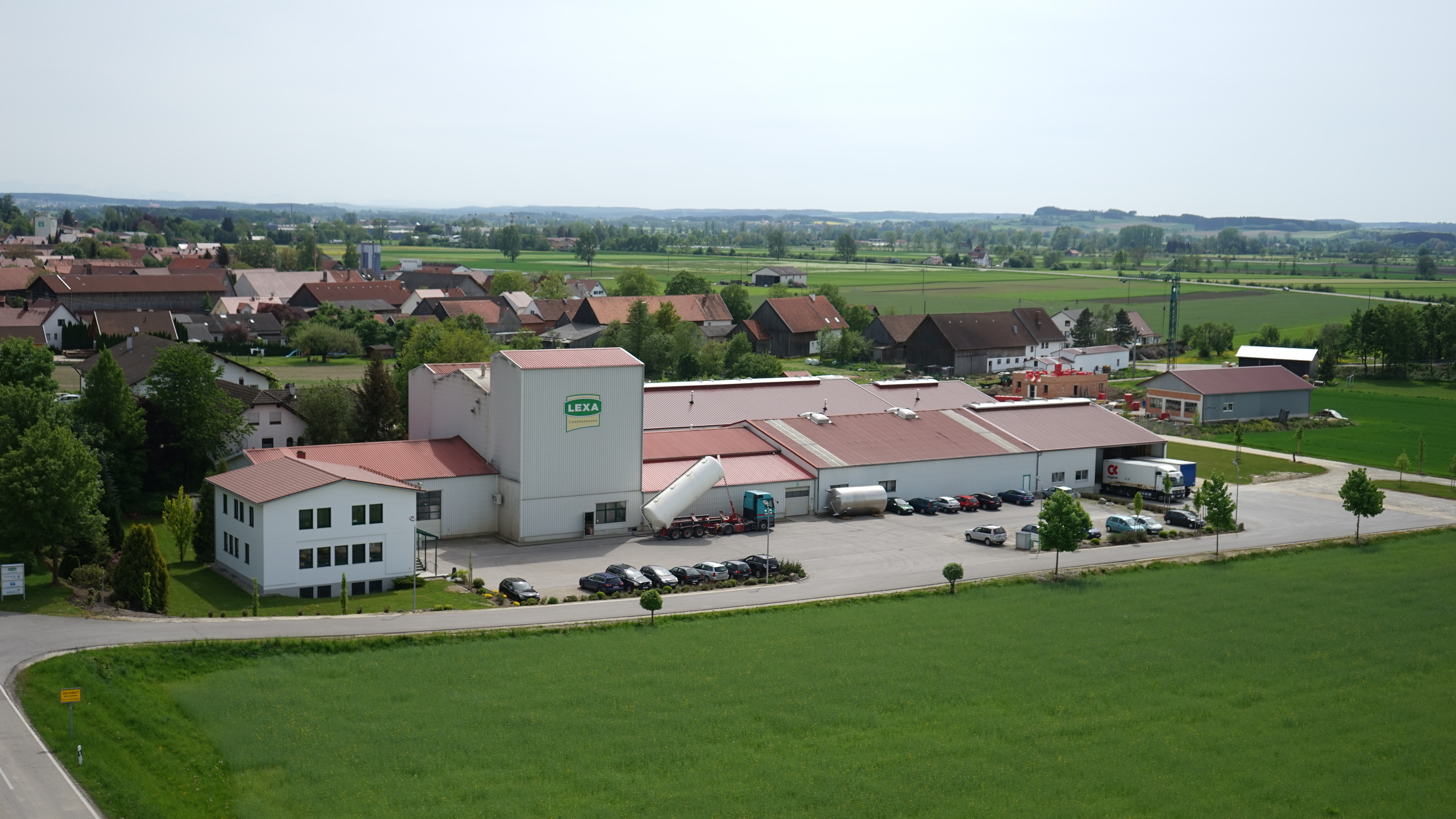 Luftaufnahme eines Lagerhauses mit Lkw und Parkplätzen, umgeben von Feldern und Wohngebäuden.