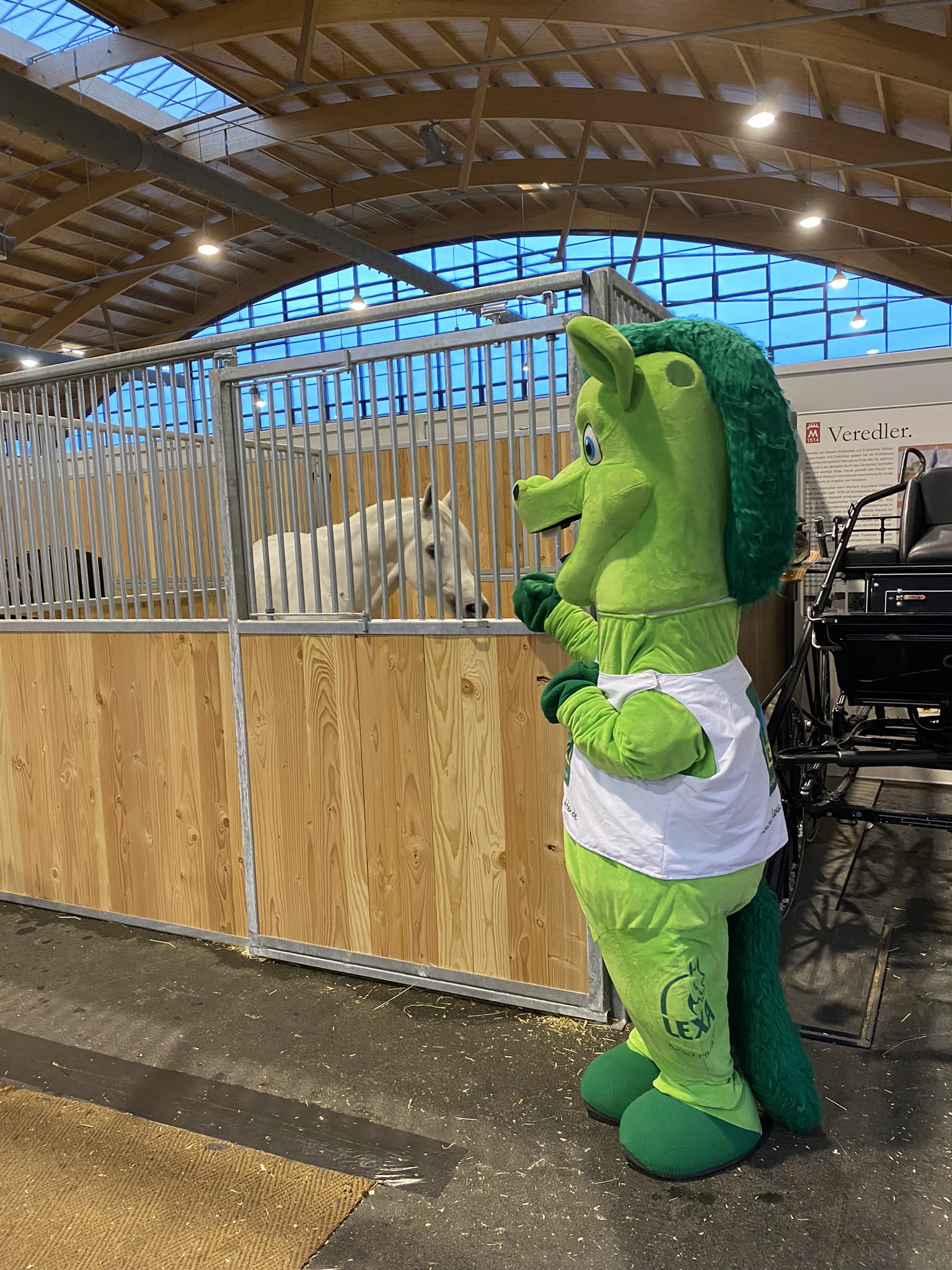 Ein grünes Maskottchen in einem Stall, das mit einem weißen Pferd interagiert.