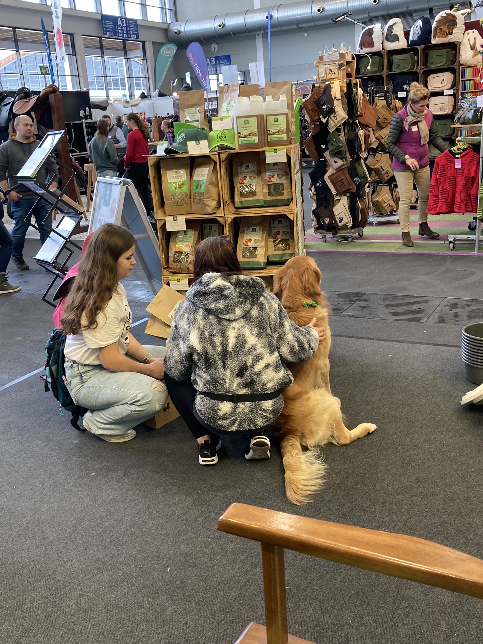 Zwei Personen knien auf dem Boden und streicheln einen Golden Retriever in einer Messehalle mit Ständen im Hintergrund.