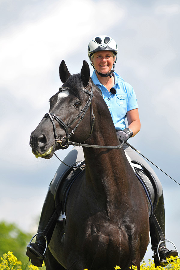 Reiterin auf einem schwarzen Pferd, lächelt in die Kamera, trägt einen Helm und ein blaues Shirt.
