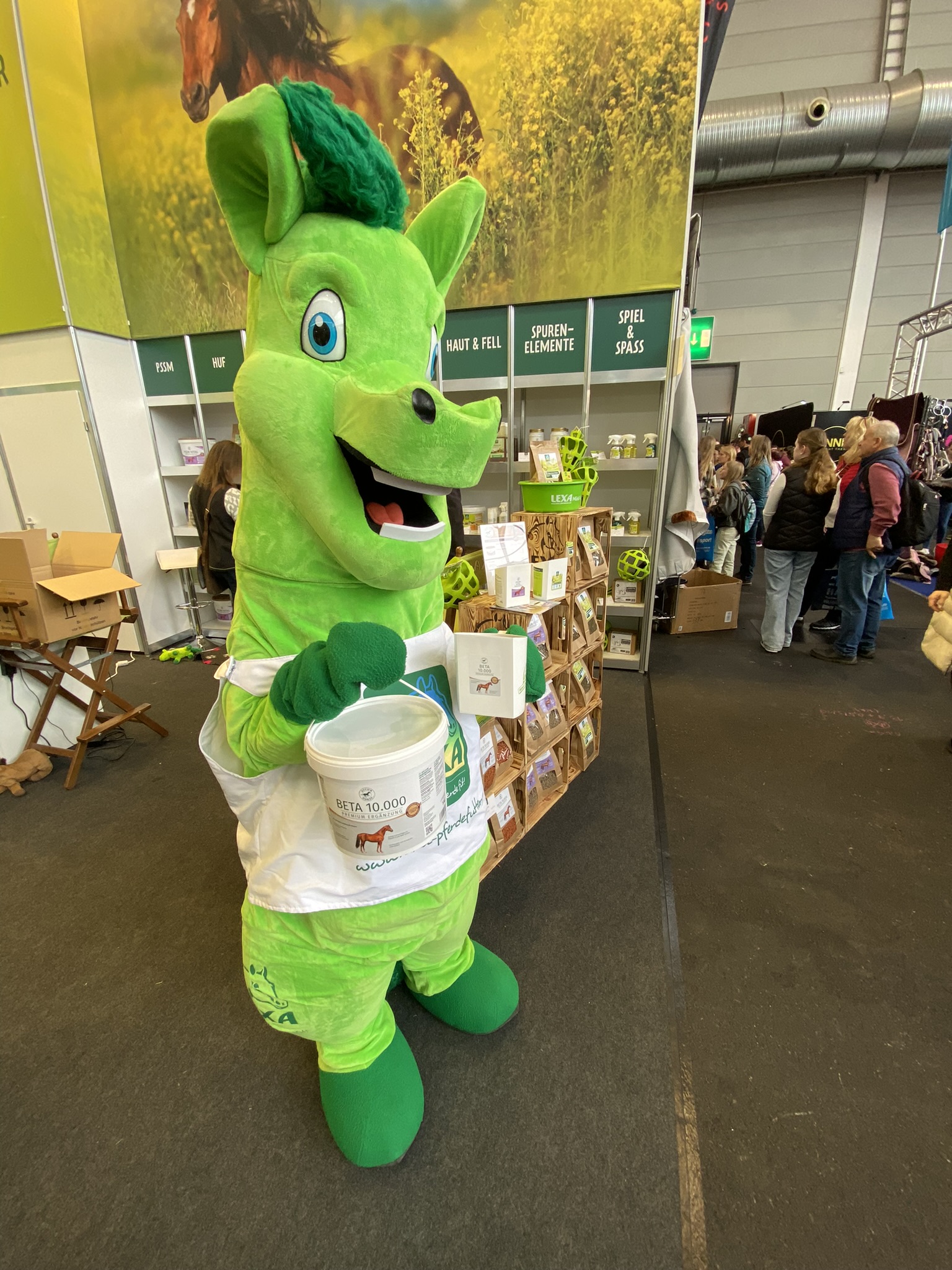 Ein grünes Maskottchen in einem Tierkostüm hält einen Eimer und steht in einer Messehalle.
